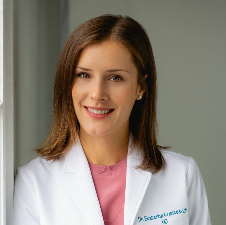 Edgemont A professional portrait of a smiling female Dr Kate in a white lab coat, standing by a window. she wears a pink shirt and has a name tag that reads "dr. ekaterina Krantsevich, ND. North Vancouver